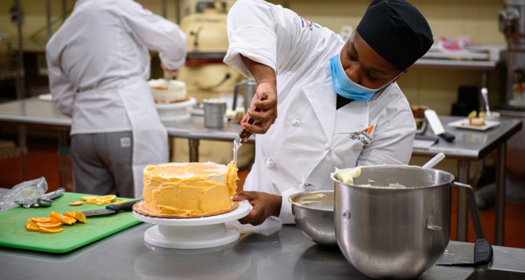 female baking student preparing a cake in a commercial kitchen with another student baker in the background