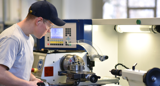 A person wearing safety glasses and a cap operates a CNC lathe machine, focusing closely on the metalwork in a well-lit workshop.