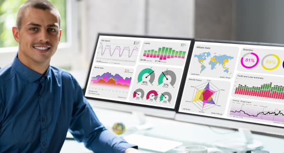 Smiling man at desk with dual monitors displaying colorful data dashboards and analytics.
