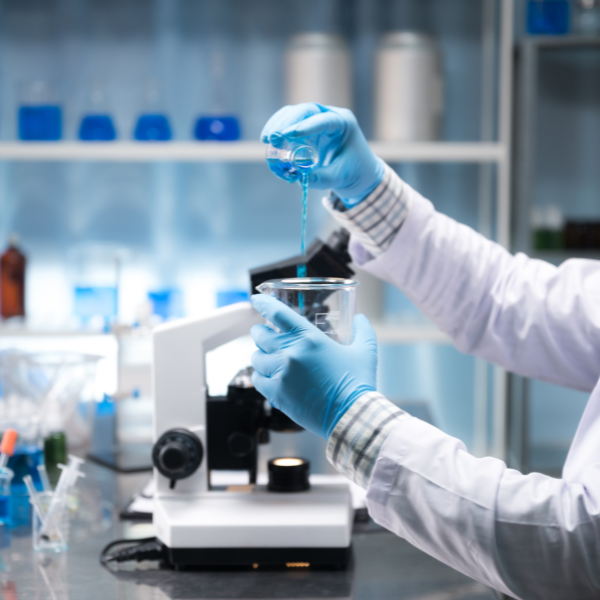 Scientist in gloves pouring blue liquid into a beaker in a lab with a microscope nearby.