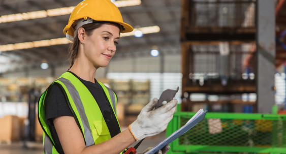 A person wearing a yellow hard hat, safety vest, and gloves inspects an item while holding a clipboard inside an industrial warehouse.