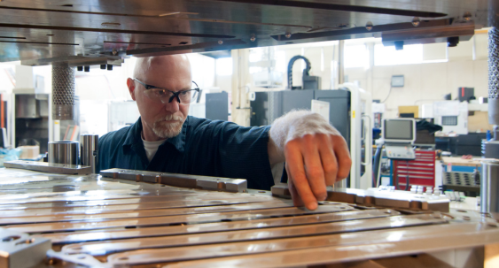 A person wearing safety glasses inspects and adjusts metal components inside a tool and die or moldmaking machine in a manufacturing workshop.