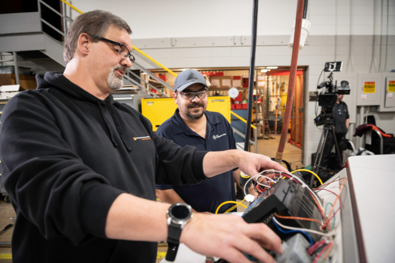 An instructor and student, both in safety glasses are gathered around a circuit board. 