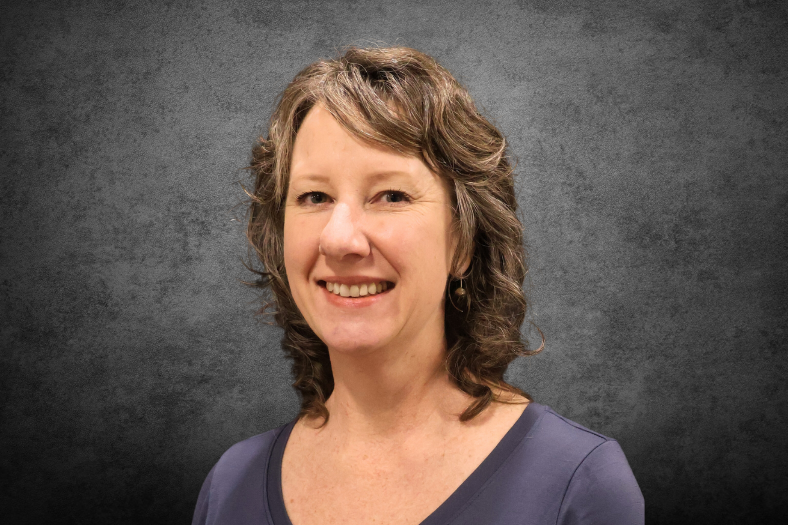 Headshot of Monica Erling with shoulder-length curly hair, smiling and wearing a purple top against a textured gray background.