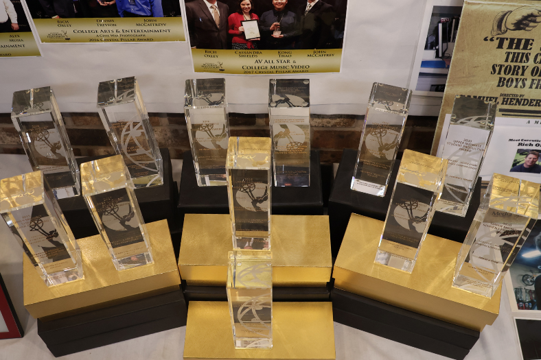 Twelve etched glass pillar awards are displayed on boxes with photos of past award winners displayed in the background.