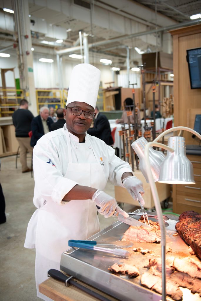 A chef in a white uniform and tall chef hat showcases his culinary arts, expertly slicing roasted meat under heat lamps in a busy commercial kitchen, with other people cooking in the background.