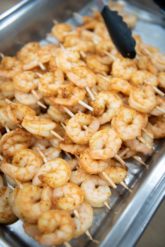 A tray filled with skewers of seasoned, cooked shrimp, demonstrating the finesse of culinary school training, with a pair of tongs resting on top.