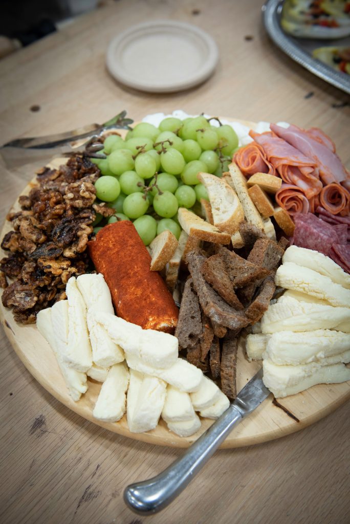 A wooden platter, showcasing the art of Culinary Arts, features green grapes, rolled cold cuts, assorted cheeses, dark and light bread slices, spiced nuts, and a cheese log with tongs and a knife on the side. A small plate is in the background.