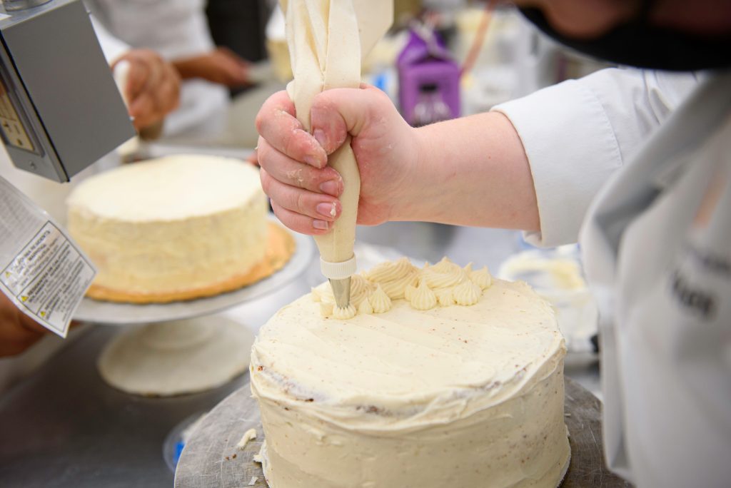 An entrepreneur in a white chef’s coat uses a piping bag to decorate a frosted cake with cream swirls. Another beautifully baked pastry sits on a stand in the background.