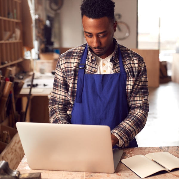 Man wearing a blue apron works on a laptop in a workshop, with a notebook open on the table beside him