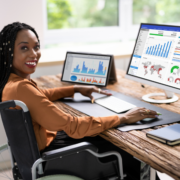 Smiling woman in a wheelchair works at a desk with data analytics on dual monitors.