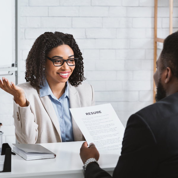 Smiling woman in a blazer speaks with a man holding a resume during a job interview at a modern office desk.