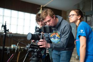 Three young people operate a professional video camera indoors, showcasing their skills in video production. One adjusts the camera while the others watch closely, appearing focused and engaged in the filming process.