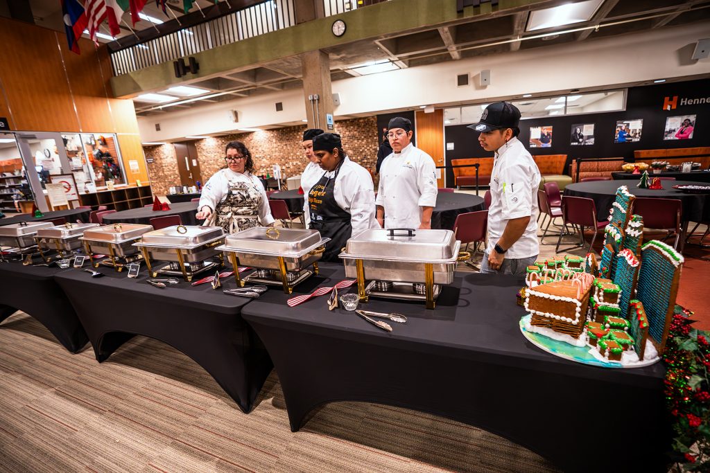 Four chefs trained in Culinary Arts stand behind a buffet table with chafing dishes and serving utensils in a spacious room. A decorated gingerbread house is displayed at the end, with round dining tables set up in the background.