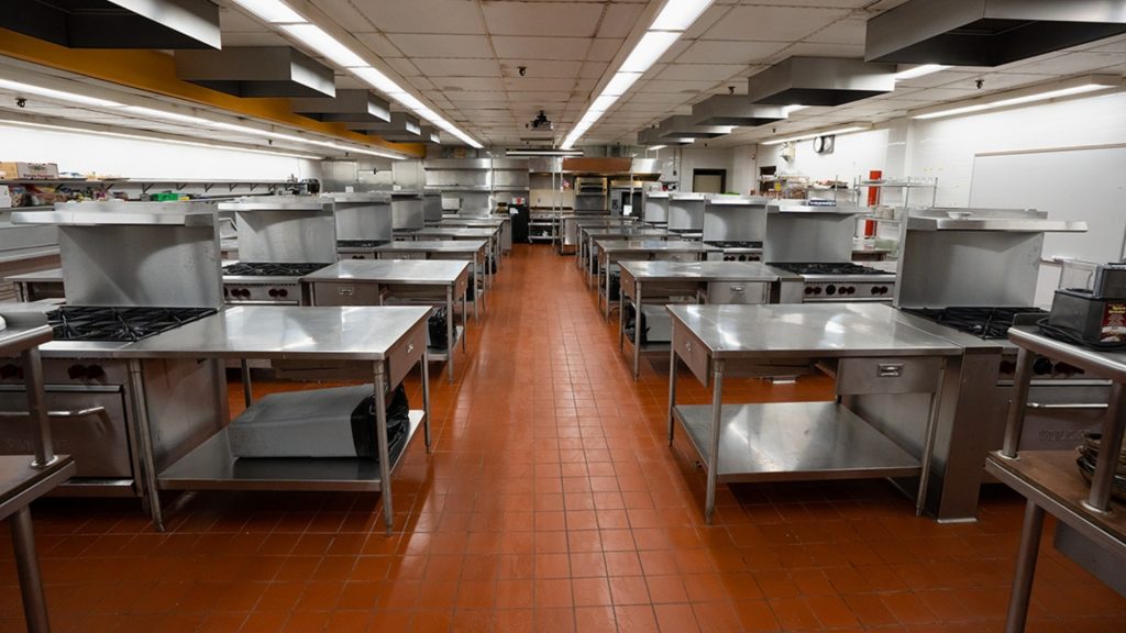 A large, industrial kitchen designed for Culinary Arts, featuring rows of stainless steel workstations and stovetops, red tile flooring, and overhead lighting. The clean, organized space is ideal for cooking classes or a bustling culinary school environment.