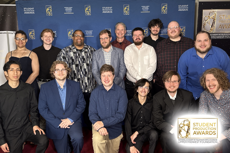 A group of fifteen students in semi-formal attire stand and kneel together, smiling for a photo at the Student Production Awards, proudly posing with a Crystal Pillar Award against an event backdrop and awards logo.